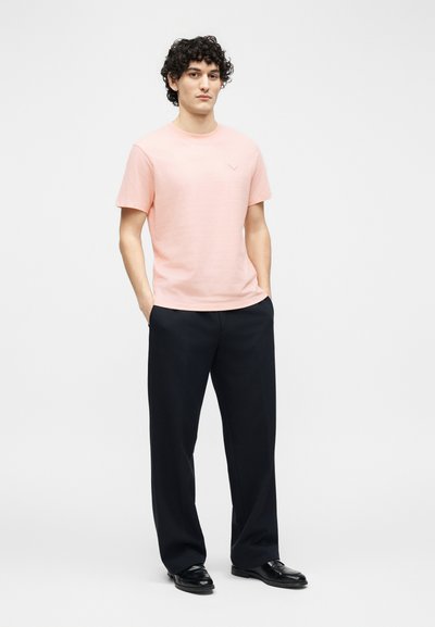 Άνδρας όρθιος με τα χέρια στις τσέπες, φορώντας ροζ ανοιχτού χρώματος κοντομάνικη μπλούζα, μαύρο παντελόνι με φαρδιά πόδια και μαύρα παπούτσια σε απλό φόντο.