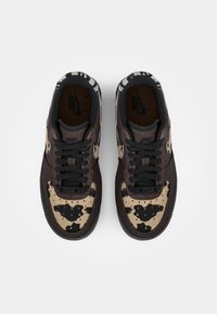 Zapatillas Nike Air con una parte superior de ante de color marrón oscuro, patrón de piel de vaca moteada en beige y negro, cordones negros y una suela de goma texturizada.