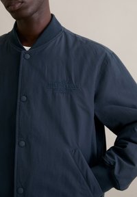 Granatowa kurtka typu bomber wykonana z gładkiej tkaniny, z zapięciem na guziki, ściągaczami na rękawach oraz haftowanym logo "Athletes Club" na piersi.