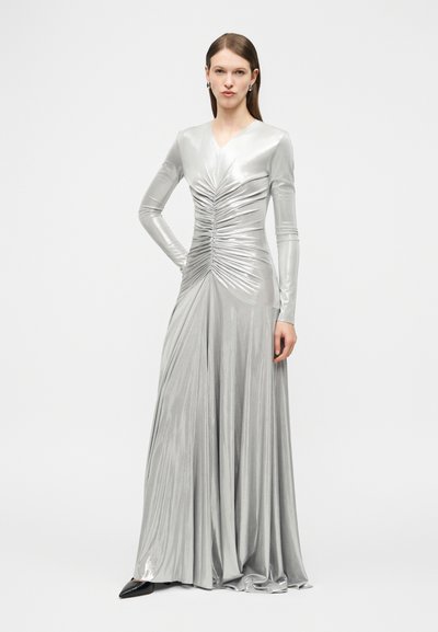 Γυναίκα με ίσια μαλλιά στέκεται φορώντας ένα μακρύ ασημένιο σατινέ φόρεμα με ρεγκλάρισμα στο μπροστινό μέρος και μαύρα μυτερά παπούτσια.