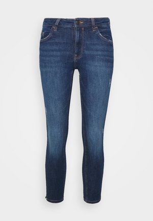 Jean skinny bleu foncé légèrement usé, avec cinq poches et une fermeture à bouton et fermeture éclair, présenté sur un fond uni.