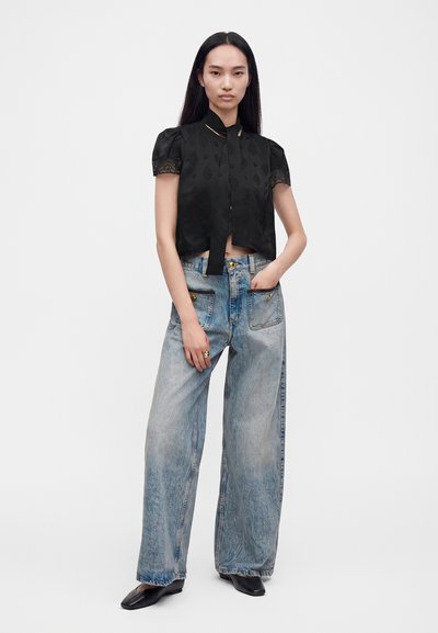 Femme debout, main dans la poche, portant un chemisier noir à manches courtes et un jean bleu délavé à jambes larges avec des chaussures noires sur fond blanc.