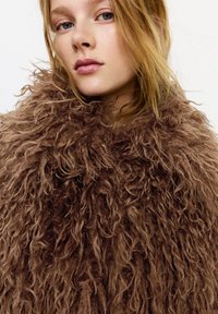 Ung kvinde med blondt hår iført en voluminøs krøllet brun pelsfrakke, der kigger let væk fra kameraet mod en ensfarvet baggrund.