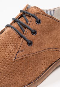 Hnědé semišové boty s perforovaným designem, s tmavými šňůrkami a očky, kontrastující s pruhovanou látkovou podšívkou a modrými švy.