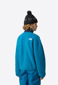 Τιρκουάζ φλις μπουφάν με ψηλό γιακά. Φέρει ένα λευκό λογότυπο στην πλάτη, φορεμένο με μαύρο πλεκτό καπέλο με φούντα και σχέδιο νιφάδας χιονιού.