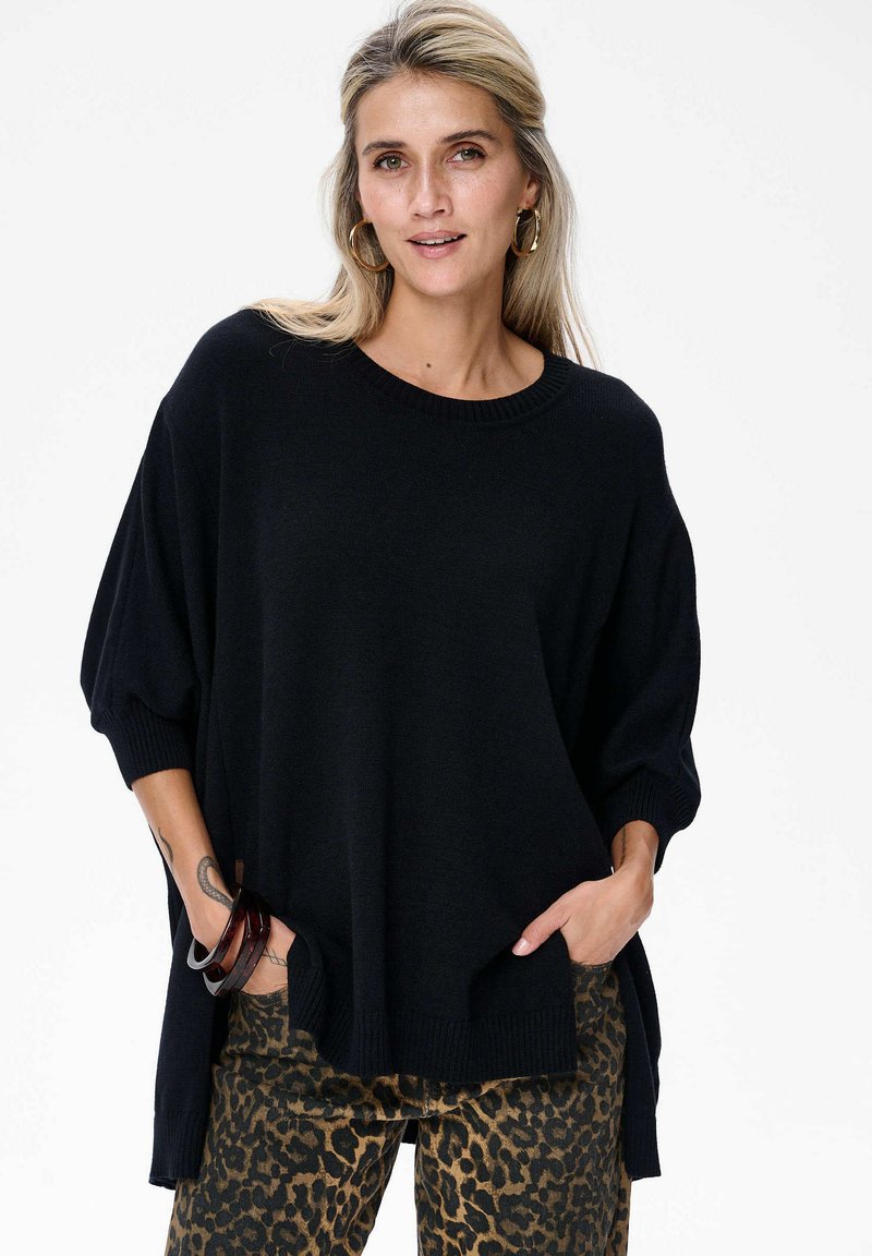 Blondynka w dużych kolczykach w kształcie kół nosi oversize'owy czarny sweter i spodnie w panterkę, ręce w kieszeniach, na tle białego tła.