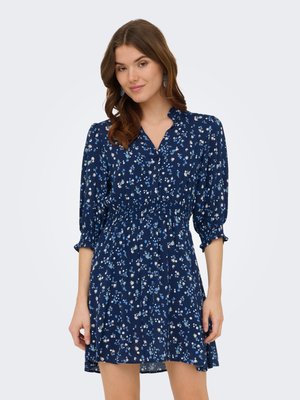 Femme portant une robe fleurie bleu marine avec des manches trois-quarts bouffantes et une taille froncée, debout devant un fond clair uni.