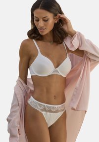 Weißer BH mit Spitzenrand und passendem Unterwäsche-Set, mit weichem Stoff und geschwungenen Formen. Das Model trägt ein hellrosa Hemd mit Knöpfen.