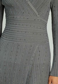 Rochie gri dintr-un material texturat cu dungi verticale, având accente metalice dispersate. Mâneci lungi și un design strâns pe talie.