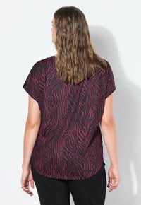 Mujer con cabello largo y ondulado de color castaño, vistiendo una camiseta de manga corta a rayas zebra en negro y rojo oscuro, de pie frente a una pared blanca, vista desde atrás.