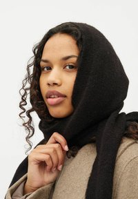 Sort uldhalstørklæde snoet om hovedet med en struktureret finish. Modellen holder en del af halstørklædet, der står i kontrast til en beige frakke.