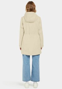 Imperméable beige avec capuche, taille cintrée et longueur mi-thigh. Doté de poches plates et d'une texture lisse. Assorti à un jean ample bleu clair.