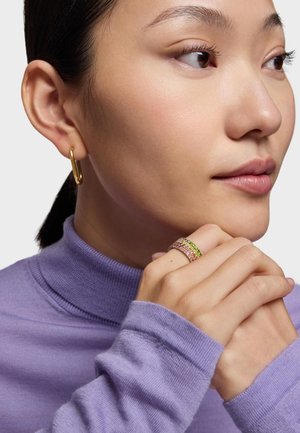 Femme en col roulé lavande, portant une boucle d'oreille en forme d'anneau doré et trois bagues ornées de pierres précieuses multicolores, mains jointes près du visage, regardant vers la droite.