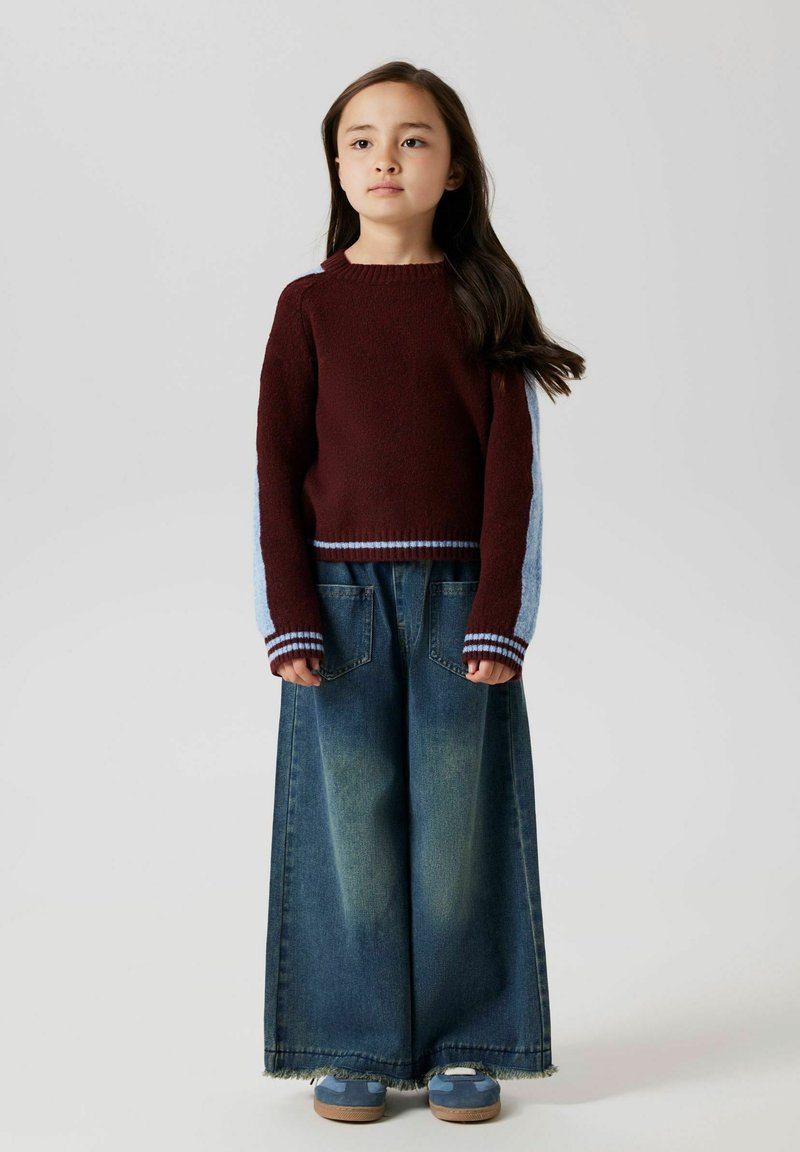 Burgundy stickad tröja med blå ärmdetaljer, i kombination med vida blå denimbyxor. Har ribbade mudd och fransdetalj vid fållen.