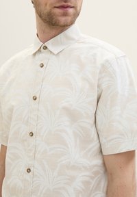 Kurzarm-Hemd mit Knöpfen in hellbeige mit weißem Palmenblattmuster, ausgestattet mit einem Standardkragen und abgerundetem Saum. Kleine braune Knöpfe.