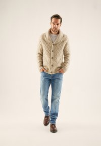 Kabeliu megztas smėlio spalvos kardiganas su šalikiniu apykakle, dideliais mygtukais ir ribomis rankogaliais, derinamas su šviesiai mėlynomis džinsinėmis kelnėmis ir rudos spalvos bateliais.