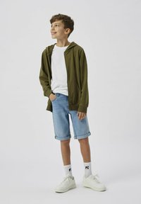 Un sweat à capuche zippé vert olive porté sur un t-shirt blanc, un short en jean bleu clair retroussé à l'ourlet, des baskets blanches, et des chaussettes blanches à motifs.