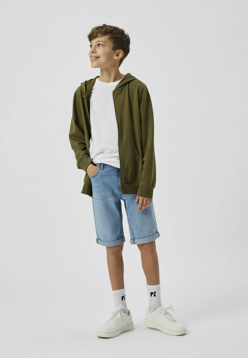 Un sweat à capuche zippé vert olive porté sur un t-shirt blanc, un short en jean bleu clair retroussé à l'ourlet, des baskets blanches, et des chaussettes blanches à motifs.