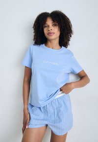 Modelka s kudrnatými vlasy má na sobě modré tričko Calvin Klein a pruhované šortky, sebevědomě pózuje na jednobarevném, světlém pozadí.