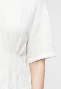 Λευκό πουκάμισο με κοντά μανίκια από υφασμάτινη υφή σε ένα μπράτσο, που δείχνει διακριτικές οριζόντιες ρυτίδες και απαλά πτυχώσεις.