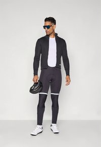 Juoda dviračių striukė su aptemptu dizainu, derinama su šviesios spalvos pirmaisiais drabužiais ir juodomis kelnėmis su baltais akcentais. Baltos dviračių batai ir šalmas.