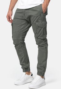 Homme portant un pantalon cargo vert olive avec des bas à fermeture éclair et des baskets blanches, debout avec une main dans la poche.