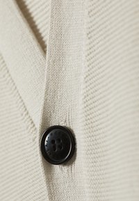 Cardigan couleur crème avec texture côtelée, doté d'un grand bouton noir gravé "WEEKDAY". Gros plan sur le bouton et le tissu.