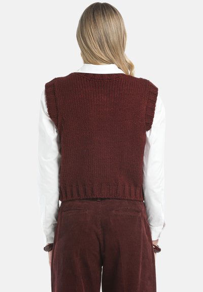 Ljus person med blont hår som bär en ärmlös stickad vinröd väst över en vit skjorta och matchande vinröda manchesterbyxor, sedd bakifrån.