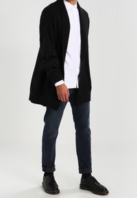 Cardigan nero in maglia con apertura frontale, maniche lunghe e tessuto testurizzato, indossato sopra una camicia bianca con bottoni e jeans scuri. Scarpe nere.