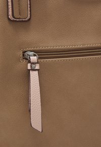 Esquina de un bolso de cuero marrón texturizado con un bolsillo con cremallera, que presenta una etiqueta de cuero rosa claro y detalles en hardware plateado.