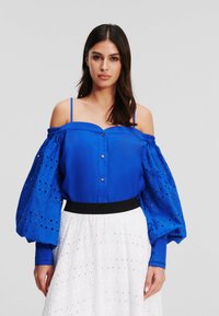 Blusa blu off-the-shoulder con maniche voluminosi, con dettagli a occhio di pernice e bottoni. Abbinata a una gonna bianca con lettering in rilievo.