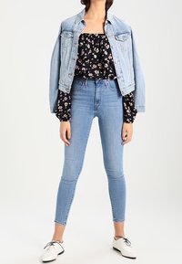 Chaqueta de mezclilla azul claro sobre una blusa negra sin hombros con estampado floral, combinada con jeans ajustados azul claro y zapatos blancos con cordones.