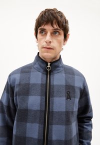 Blauwe en donkergrijze geruite zip-up jas met een hoge kraag, gemaakt van zachte stof. Heeft een geborduurd logo en een zilveren rits.