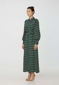 Robe maxi de couleur vert foncé avec un motif géométrique de carrés roses et blancs, dotée d'un col, de manches longues et d'une taille ceinturée.