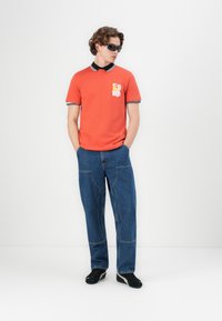 Orange polo skjorte med sort krave og manchetter; hvid grafisk detalje på brystet. Båret med løse blå denim jeans og sorte sneakers.