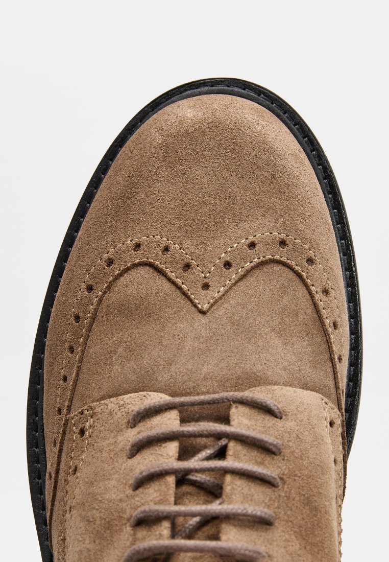 Brązowe zamszowe buty typu wingtip z okrągłym noskiem, ozdobnymi perforacjami i ciemnymi sznurowadłami. Czarna gumowa podeszwa z teksturowanym wykończeniem.
