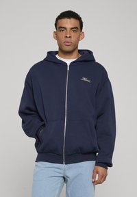 Felpa con zip di colore blu navy realizzata in un mix di cotone, dotata di tasca frontale, cappuccio con cordino e testo ricamato in oro sul lato sinistro del petto.