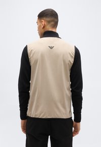 Άνδρας που φοράει μπεζ χωρίς μανίκια γιλέκο με μαύρο γιακά και μαύρο πουκάμισο με μακριά μανίκια, στέκεται γυρισμένος με την πλάτη του σε απλό φόντο.