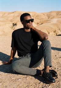 Homme portant un t-shirt noir, un pantalon gris et des lunettes de soleil assis sur un sol rocheux dans un paysage désertique avec des collines couleur sable.