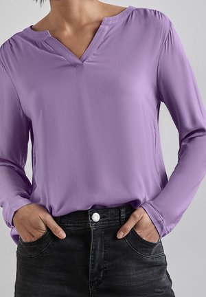 Persoon draagt een lavendelkleurige blouse met lange mouwen en V-hals, ingestopt in zwarte hooggetailleerde spijkerbroek, met handen in de zakken.