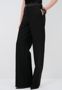 Pantalons larges noirs en tissu lisse, à taille haute, avec des poches avant et une silhouette ajustée et épurée. Pas de motifs ni d'ornements visibles.