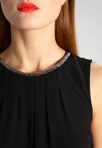 Femme portant un haut sans manches noir avec un col en perles argentées, du rouge à lèvres rouge et des cheveux raides brun-roux, sur fond gris.