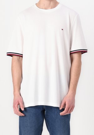 White short-sleeve t-shirt with red, white, and navy striped cuffs and small logo on the chest, worn with blue jeans.