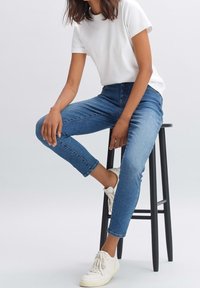 Femme portant un t-shirt blanc, un jean bleu et des baskets blanches, assise sur un tabouret noir sur un fond uni.