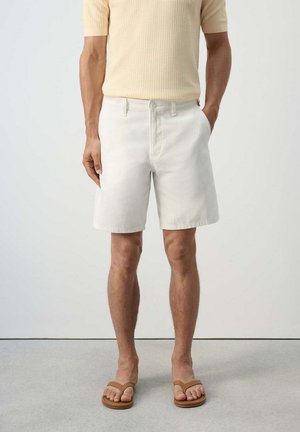 Mann trägt weiße knielange Shorts, hellbeige strukturierte Kurzarmshirt und braune Flip-Flop-Sandalen, steht vor einem einfarbigen Hintergrund.