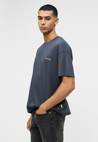 Νεαρός άνδρας με κοντά μαλλιά, που φοράει άνετο σκούρο γκρι T-shirt και μαύρο τζιν, στέκεται με το ένα χέρι στην τσέπη μπροστά σε απλό φόντο.