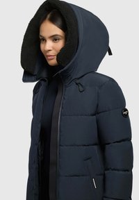 Puffjakke i marineblå med en stor, sort fleeceforet hætte. Har lynlåse og et logo patch på venstre ærme. Struktureret materiale.