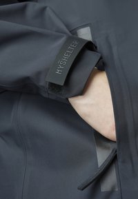 Tamno siva vodootporna jakna glatke teksture, s čičak trakama označenim "MOJE SHRANJENJE" i otvorom za bočnu džep.