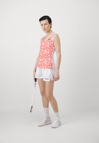 Pink and white polka dot athletic tank top with a scoop neckline, paired with white shorts. Wields a tennis racket, wearing white sneakers.