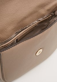 Vaalea nahkainen käsilaukku, jossa on sileä pinta, kultasävyisellä ympyränäppäyksellä ja sisällä brändilapulla varustettu tasku.